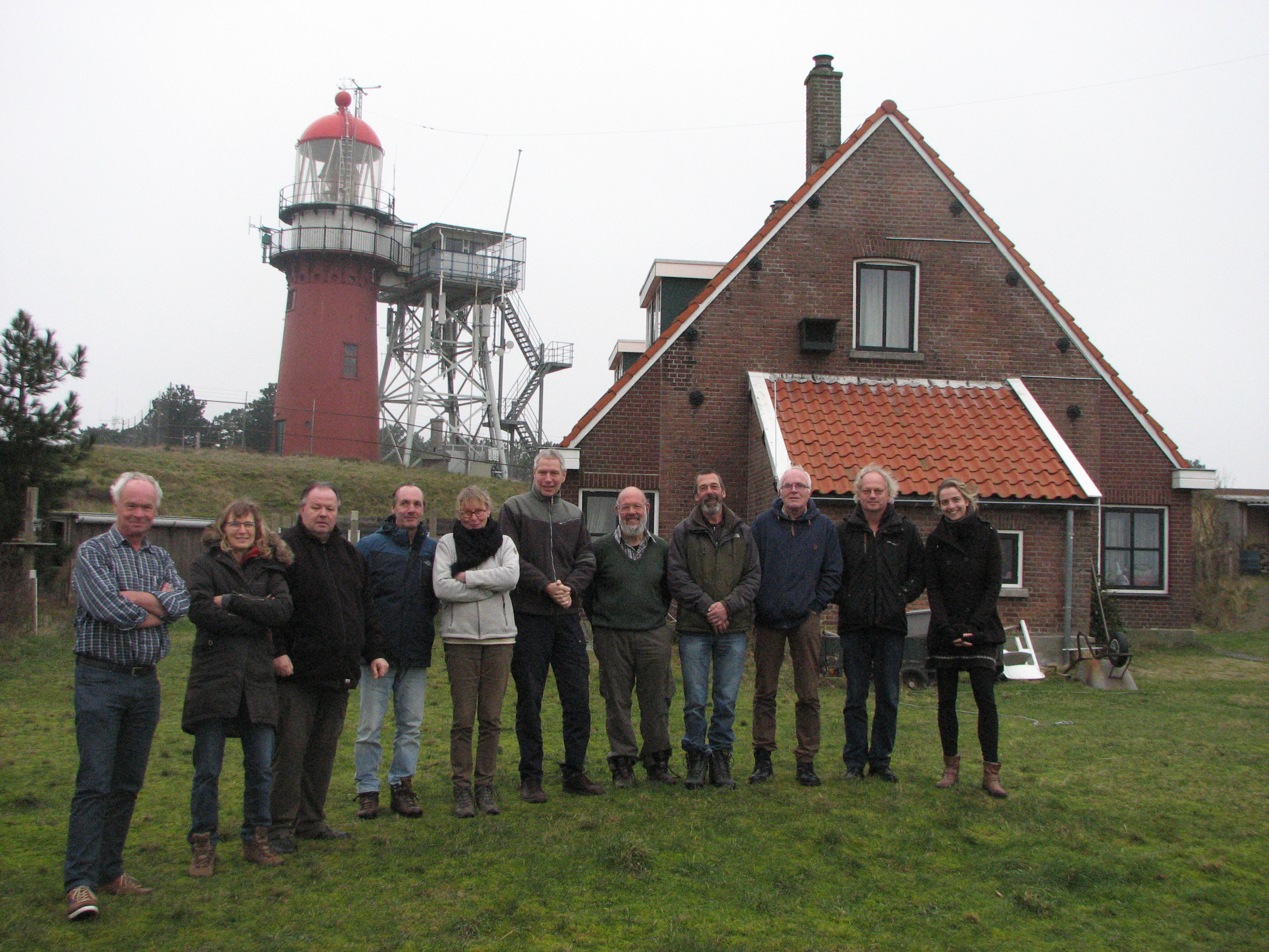 Eleven people posing in front of a stone house. A red lighthouse in the background.